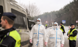 Zdjęcie przedstawia funkcjonariuszy Policji oraz sanitariuszy w kombinezonach.