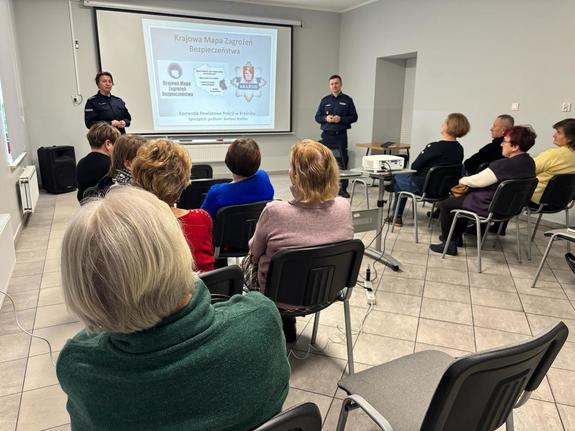 Zastępca Komendanta Powiatowego Policji w Kraśniku oraz policjantka wydziału prewencji stoją przy tablicy multimedialnej przed gronem osób siedzących na krzesłach.jpeg