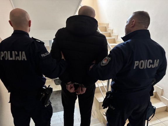 trzech mężczyzn stoi przed schodami, od lewej policjant pośrodku zatrzymany z rękami skutymi kajdankami z tyłu i po prawej stronie stoi policjant