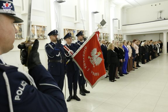 po lewo widać policjant z szablą oraz sztandar policyjny wraz z policjantami a także pracownicy cywilni stojący po prawo