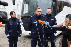 policjanci stojący przy mikrofonach przed autokarem koloru białego