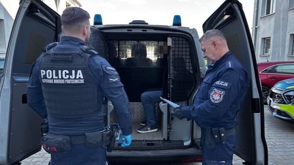 zatrzymany siedzi w radiowozie, przed radiowozem stoi dwóch policjantów, jeden tyłem drugi bokiem, sprawdzając dokumenty