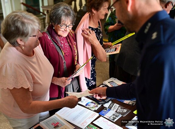 seniorki odbierają ulotki od policjanta przy stoisku profilaktycznym
