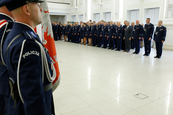 z lewej strony widać napis policja na rękawie munduru policjanta a w oddali stoi kadra kierownicza w mundurach policyjnych